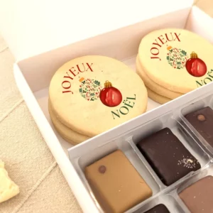 Gros plan sur biscuits “Joyeux Noël” avec chocolats dans un coffret blanc aux détails dorés.