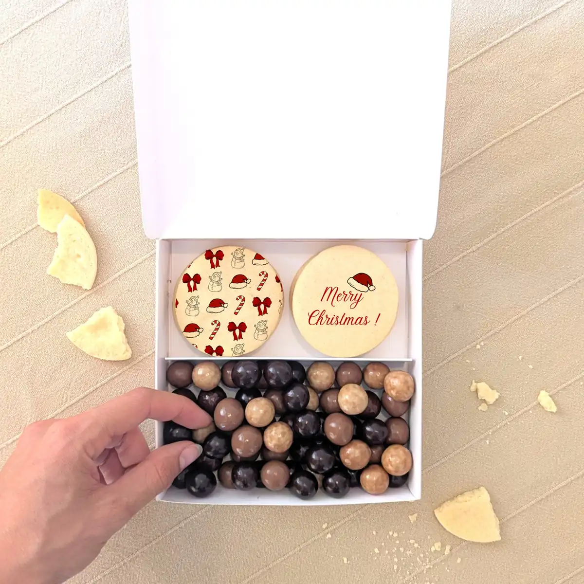 biscuis personnalisés dans coffret gourmand de noel Coffret gourmand de Noël avec deux biscuits personnalisés “Merry Christmas” et assortiment de chocolats en boîte blanche à reliefs dorés.